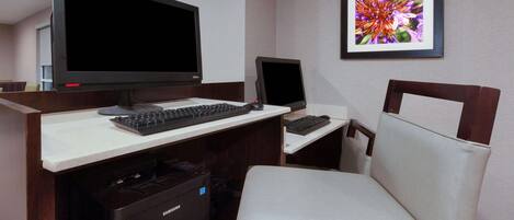A hotel room with a desk, two monitors, a keyboard, a Samsung dishwasher, and a framed floral artwork on the wall.