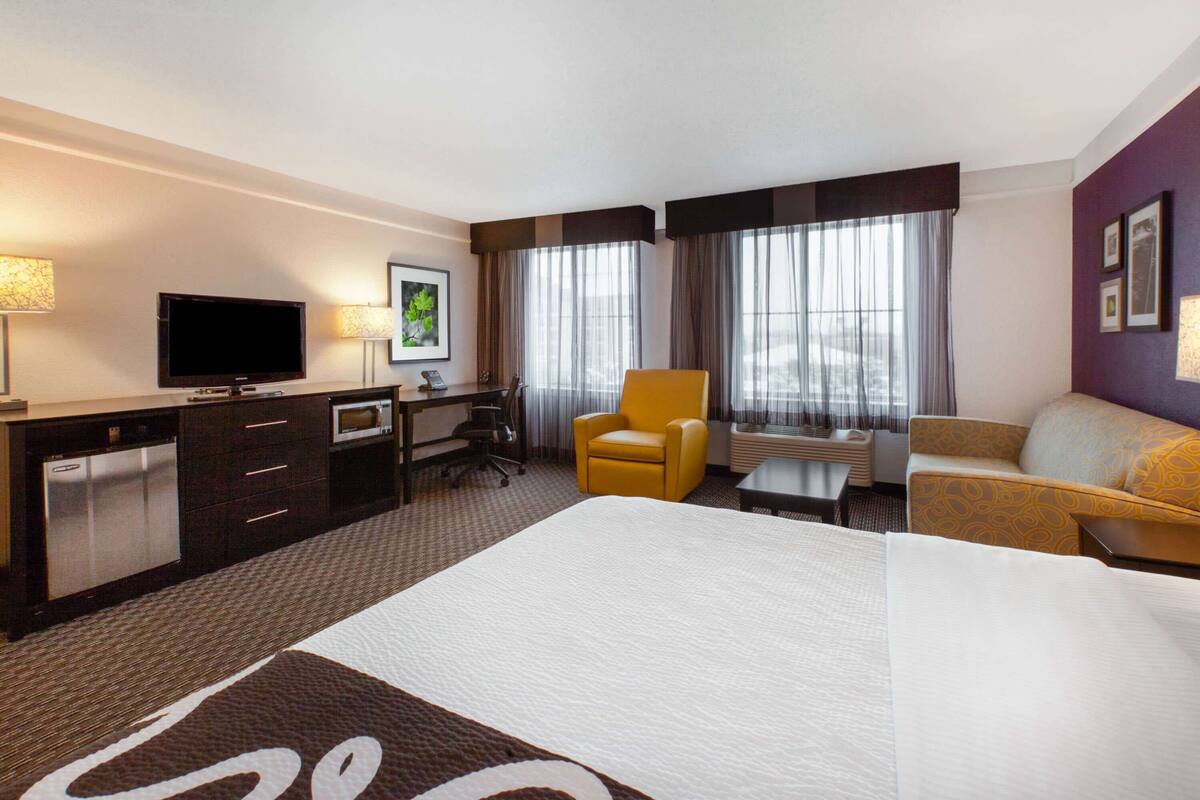 A hotel room with a large bed, a desk with a chair, a television, a minibar, and a sofa.