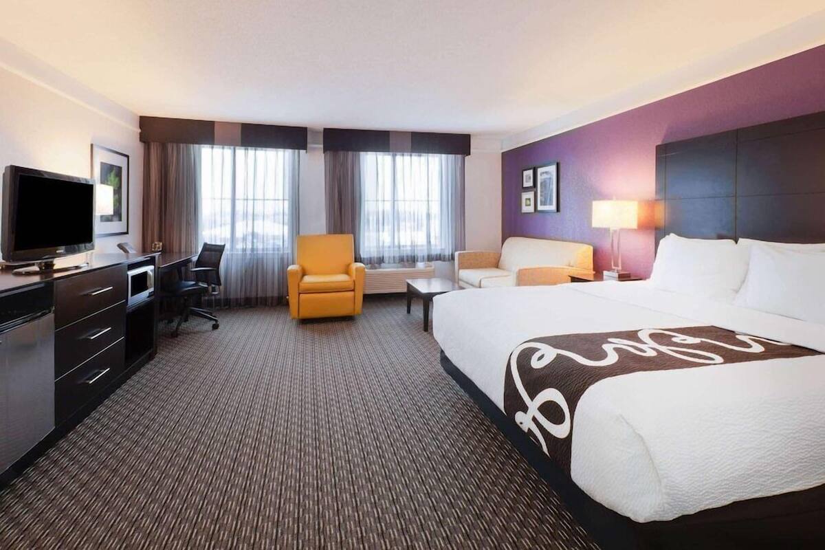 A hotel room with a large bed, a desk with a TV, a chair, a sofa, and a window with curtains.