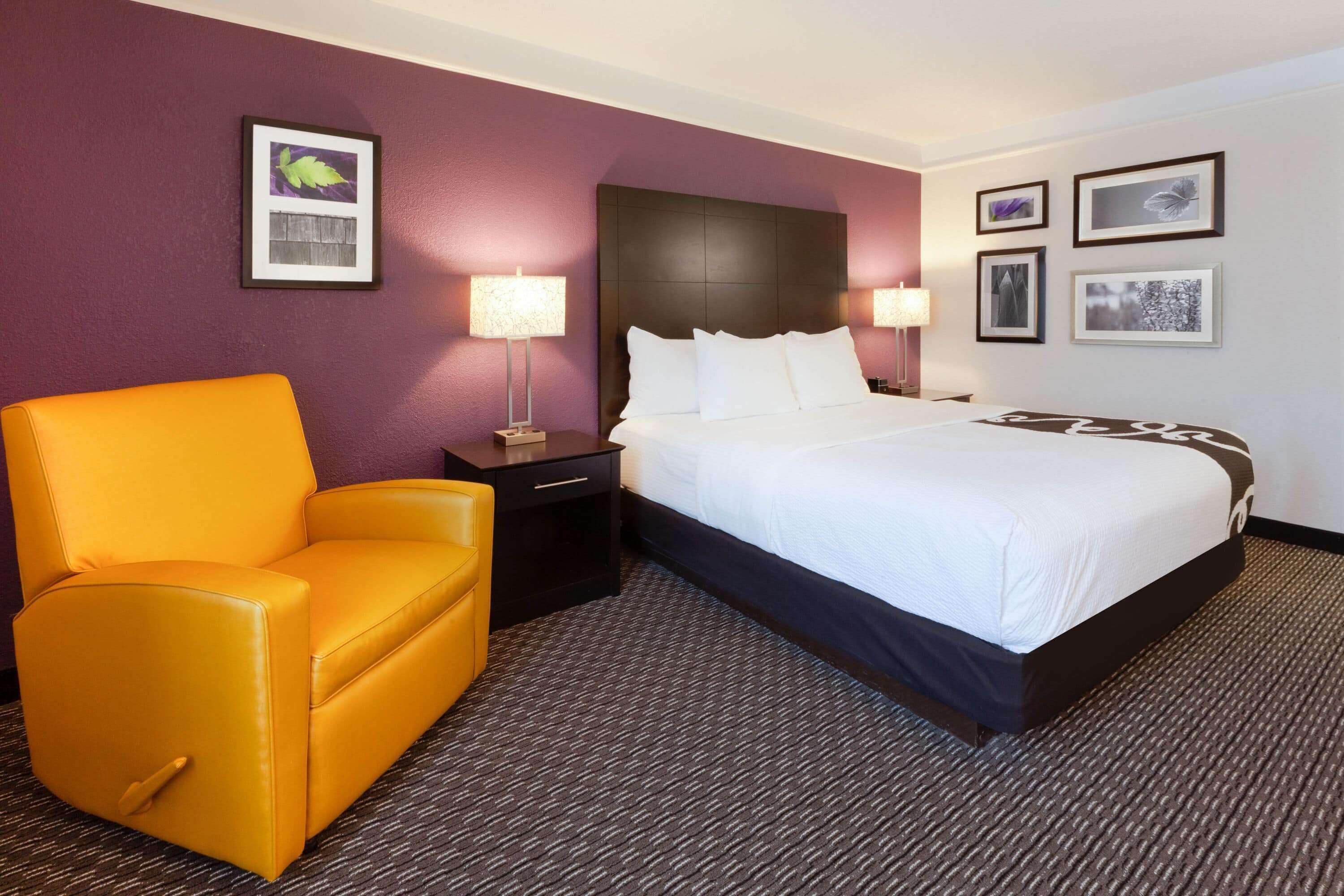 A hotel room with a large bed, a yellow armchair, a nightstand with a lamp, and framed pictures on the wall.