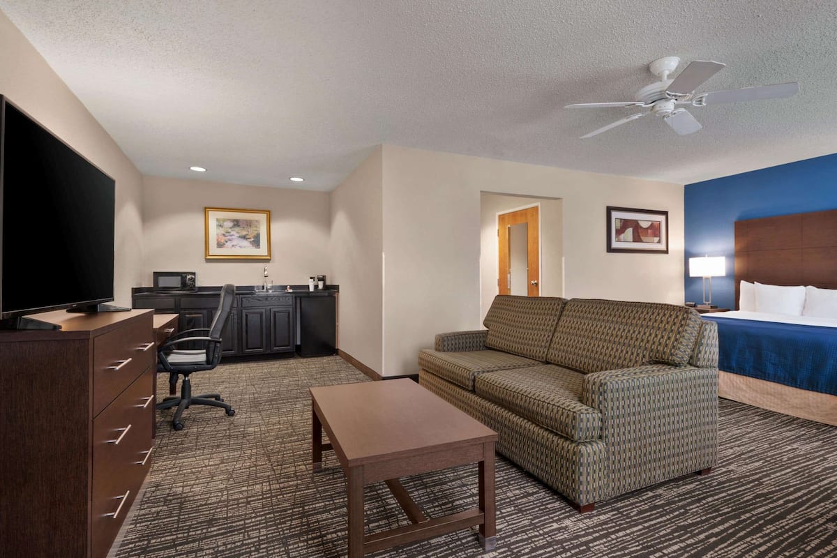 Ein Hotelzimmer mit Sofa, Fernseher, Schreibtisch, Stuhl, kleinem Tisch, Deckenventilator und Bett.