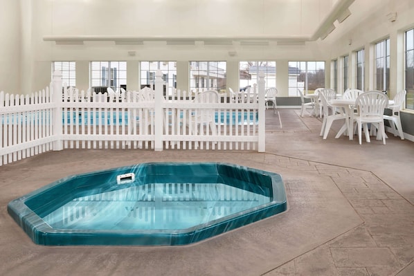 A large indoor swimming pool with a white fence, surrounded by white plastic chairs and tables, with large windows in the background.