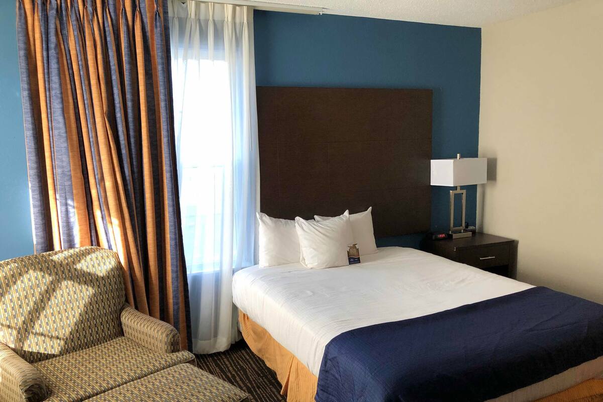 Ein Hotelzimmer mit Bett, Sessel, Nachttisch und einem Fenster mit Vorhängen.