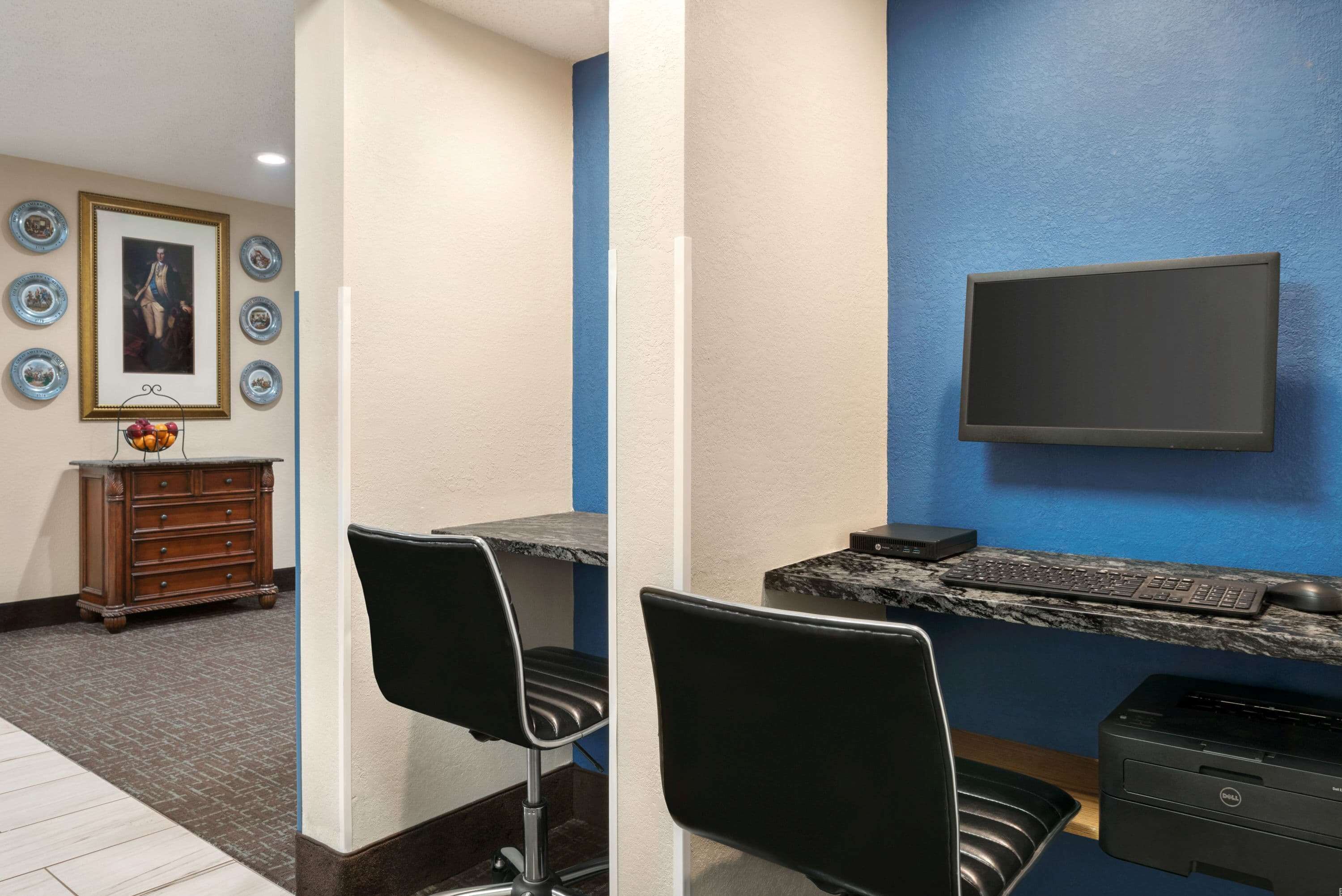 A hotel room with a desk, two chairs, a flat-screen TV mounted on the wall, a printer, and a decorative wall with framed pictures and a fruit bowl.