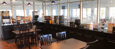 Un restaurante con una decoración moderna, que cuenta con mesas y sillas de madera, una barra de café y grandes ventanales que permiten la entrada de luz natural.