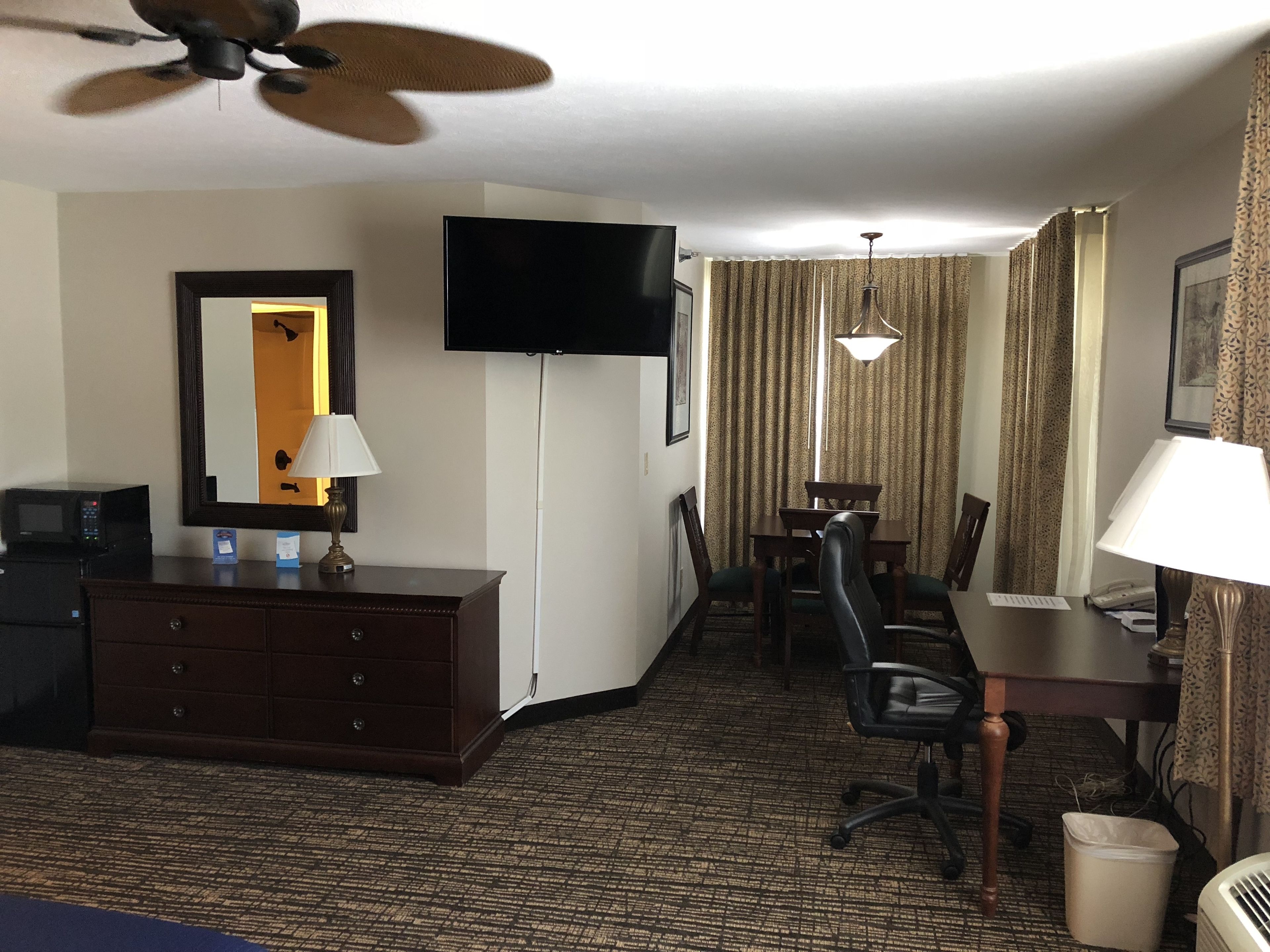 A hotel room with a flat-screen TV, a desk with a chair, a dining table with chairs, a ceiling fan, and a large mirror.