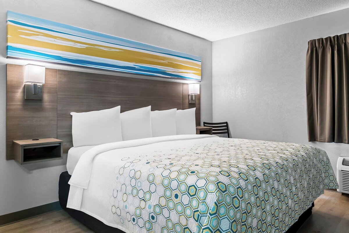 A modern hotel room with a large bed, a headboard with built-in lighting, a bedside table, and a wall-mounted artwork.