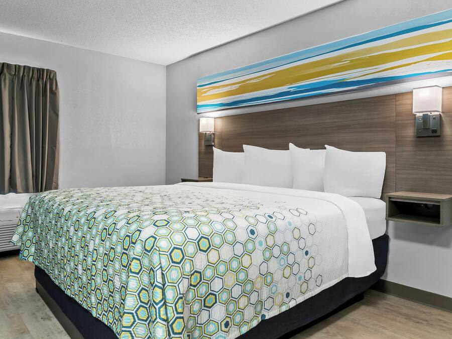 A modern hotel room with a large bed, a headboard with built-in lighting, a bedside table, and a wall-mounted air conditioning unit.