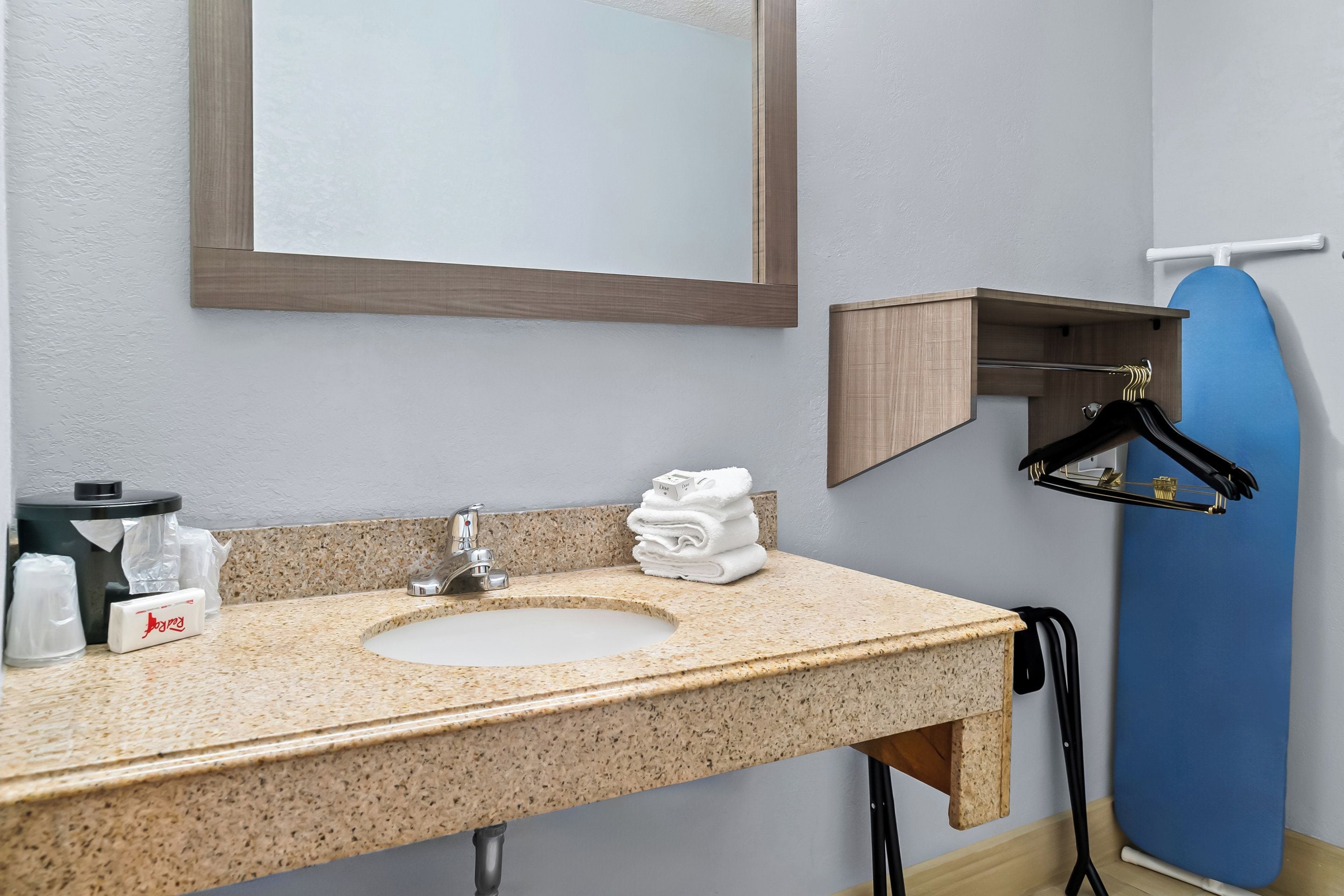 A bathroom with a granite countertop sink, a mirror, a towel rack, and a blue ironing board.