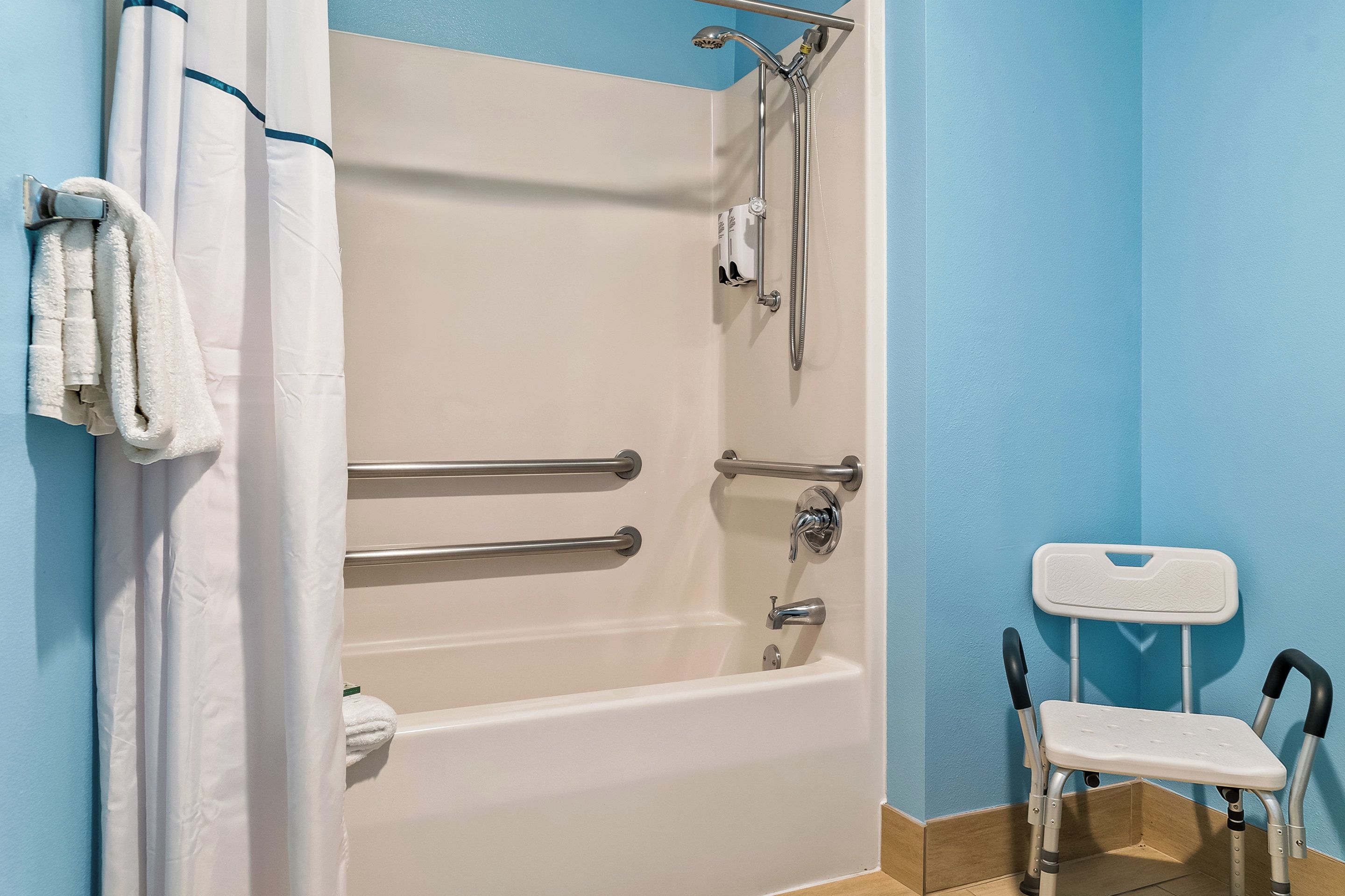 Une salle de bain avec une baignoire blanche, des barres d’appui, une pomme de douche et une chaise blanche.