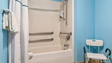 Une salle de bain avec une baignoire blanche, des barres d’appui, une pomme de douche et une chaise blanche.