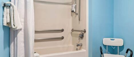 Un baño con una bañera blanca, barras de apoyo, un cabezal de ducha y una silla blanca.