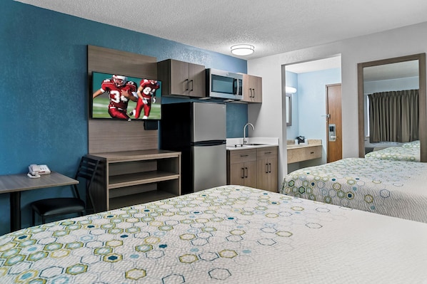 A hotel room with two beds, a TV mounted on the wall, a microwave, a refrigerator, a desk with a chair, and a small bathroom visible through an open door. - HomeTowne Studios by Red Roof Flint (Flint)