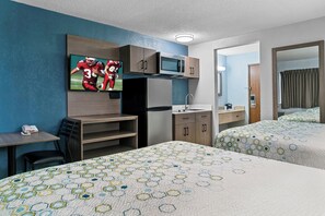 Habitación de hotel con dos camas, televisor montado en la pared, microondas, refrigerador, escritorio con silla y un baño pequeño visible a través de una puerta abierta.