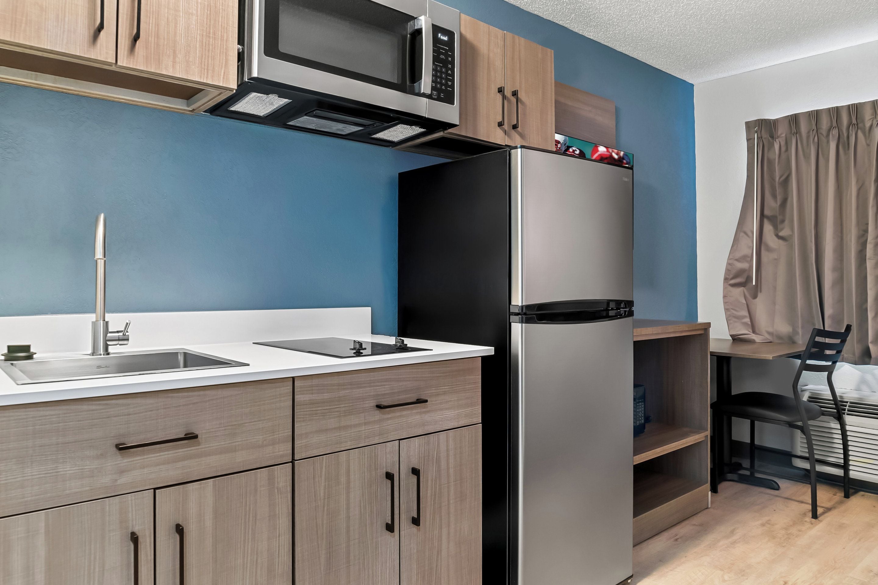 A modern kitchen with a stainless steel refrigerator, a microwave, and a sink. There is a wooden cabinet above the microwave and a chair with a table next to the kitchen.