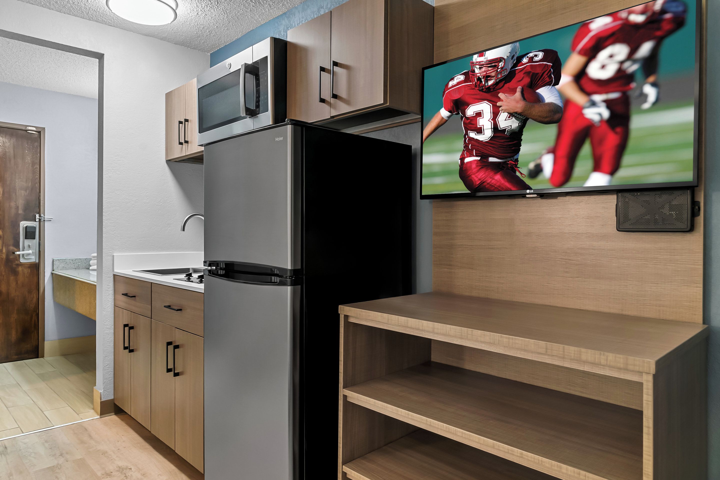 A modern kitchen with a built-in refrigerator, wooden cabinets, a microwave, and a flat-screen TV displaying a football game.