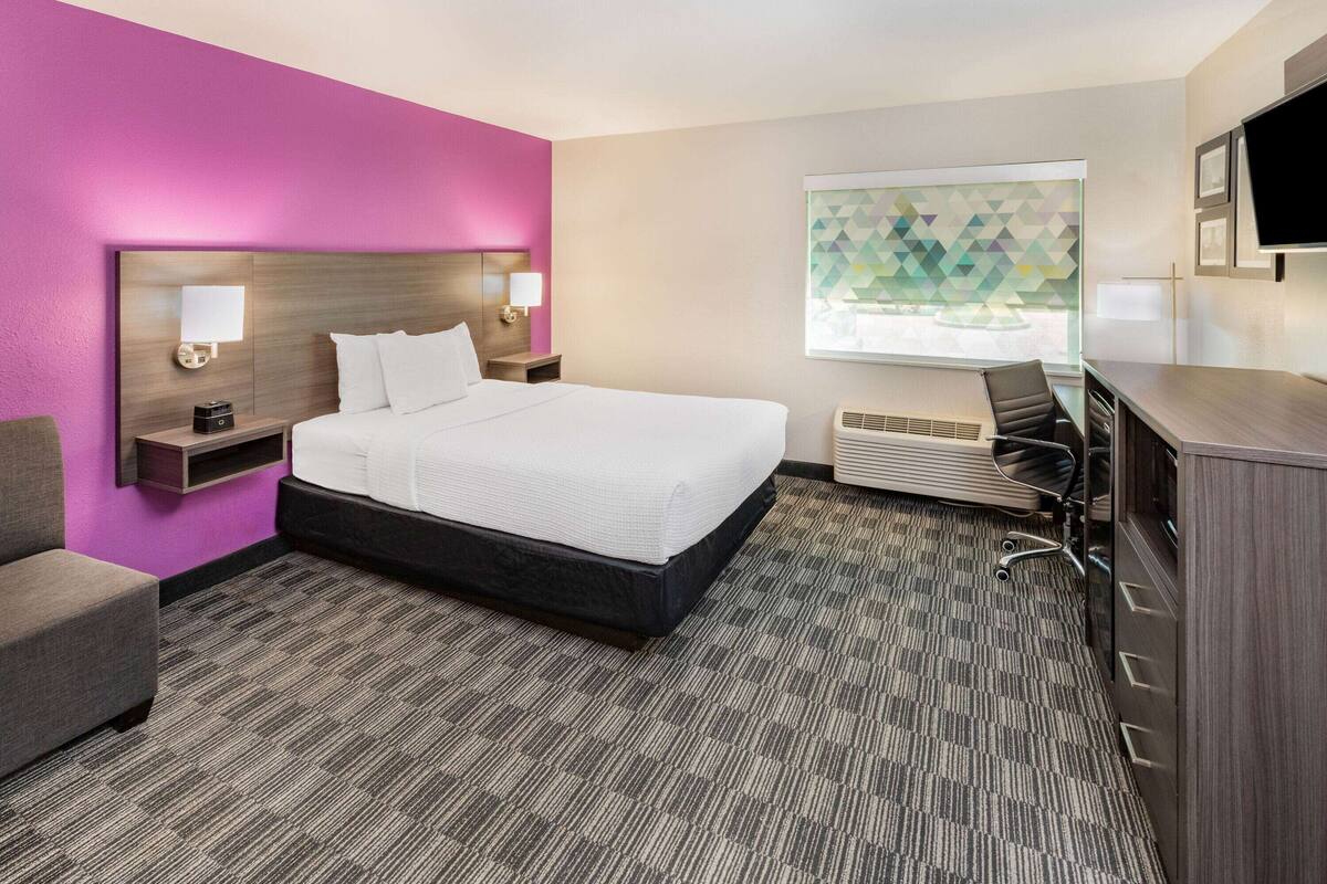 Ein Hotelzimmer mit einem großen Bett, einem Schreibtisch, einem Sessel, einem Fernseher und einem Fenster mit geometrischer Aussicht.