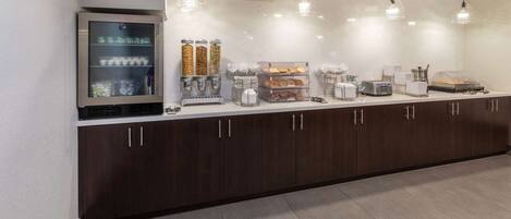 Una barra de desayuno moderna con un refrigerador, máquinas de café y una variedad de alimentos.