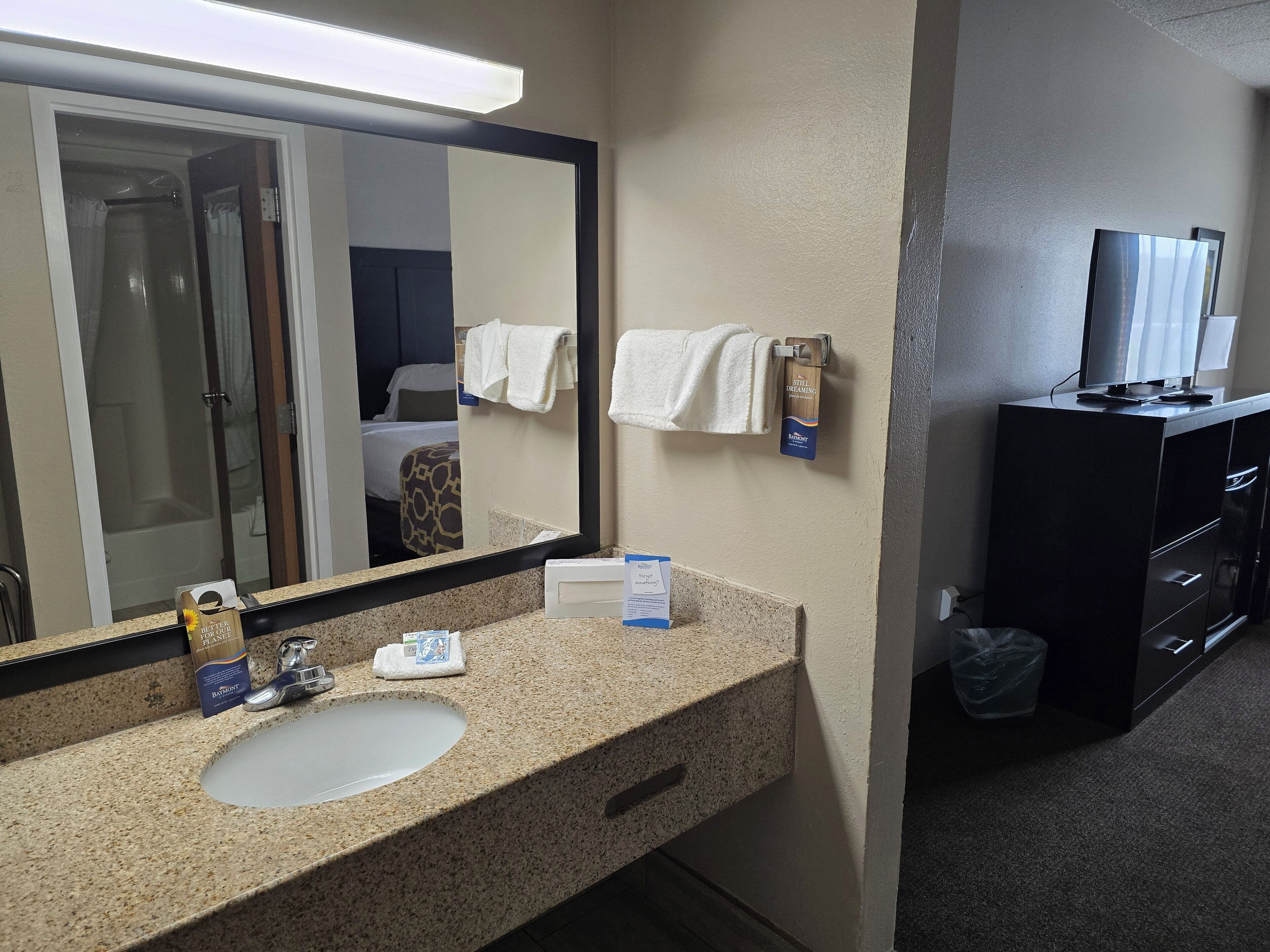 Un baño de hotel con lavamanos de encimera de granito, un espejo grande, un perchero, un televisor sobre un mueble y un cubo de basura.