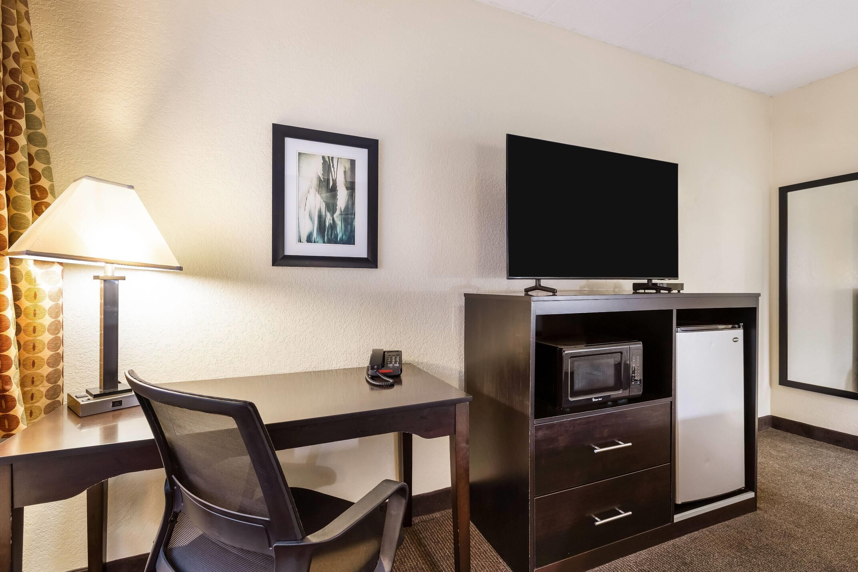 A hotel room with a desk, chair, TV, microwave, refrigerator, and a framed picture on the wall.