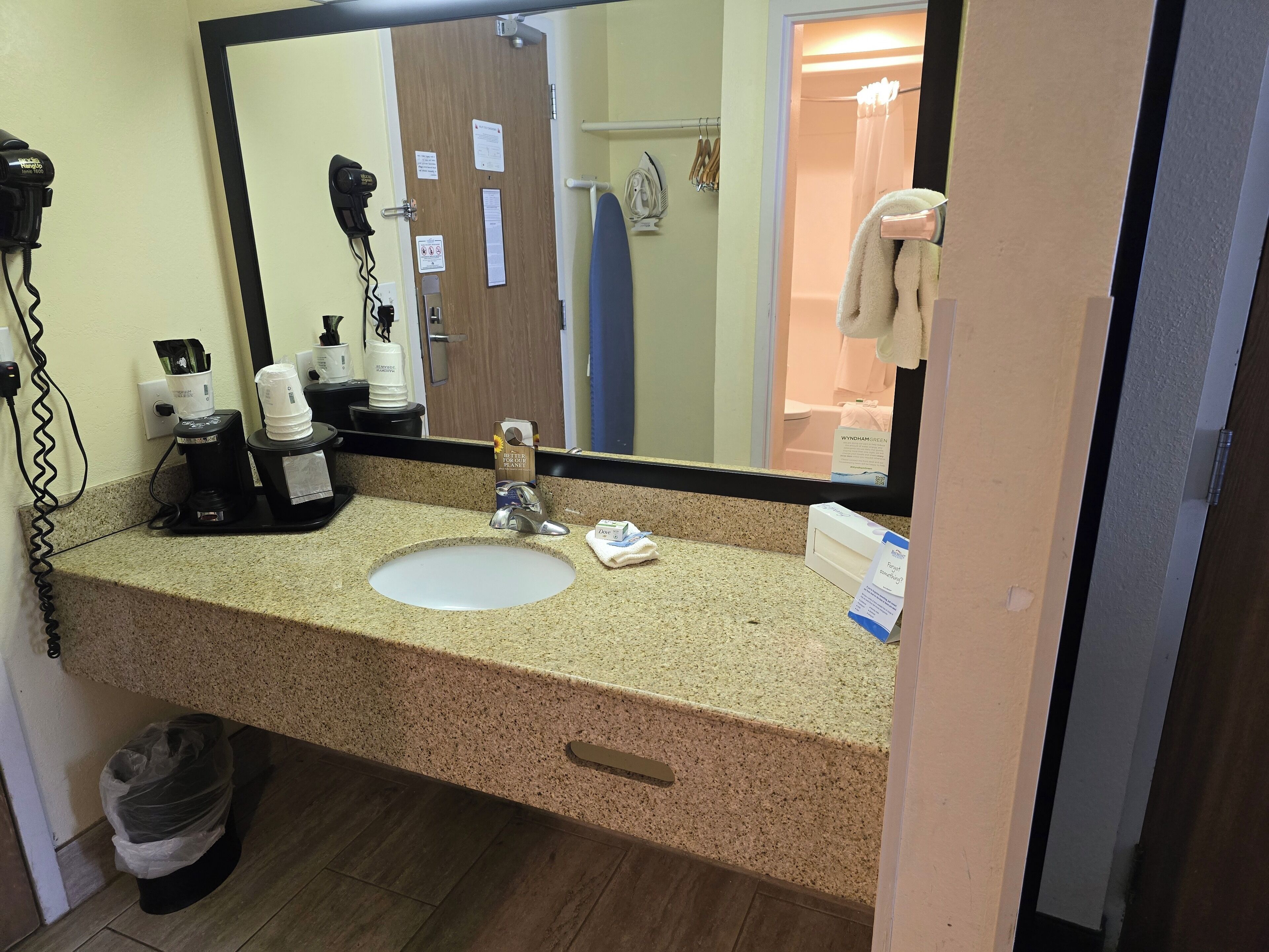 Baño de hotel con encimera de granito, un lavamanos, secador de pelo, plancha y cubo de basura.