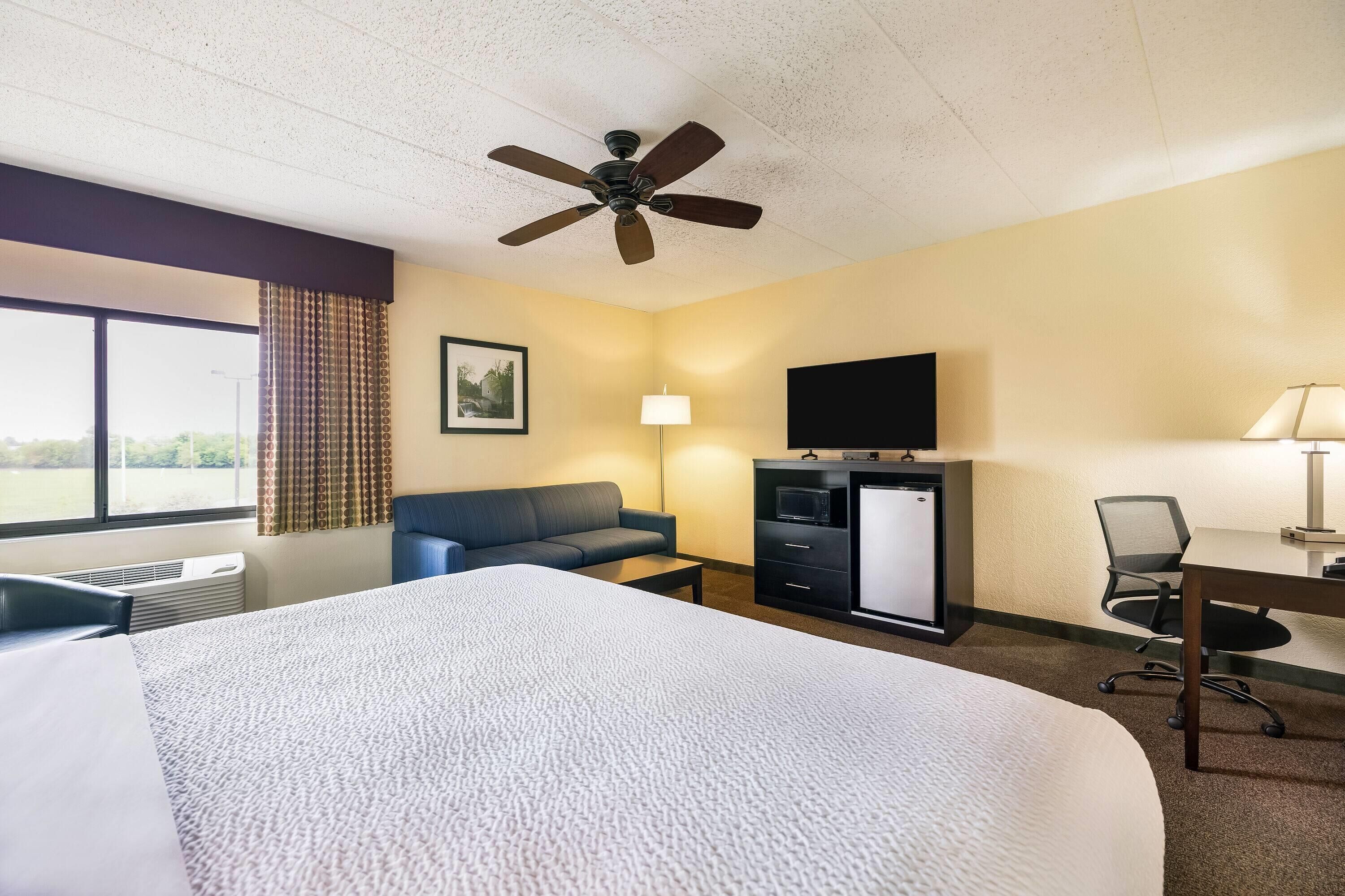 Quarto de hotel com cama, escrivaninha, cadeira, televisão, ventoinha de teto e janela com cortinas.