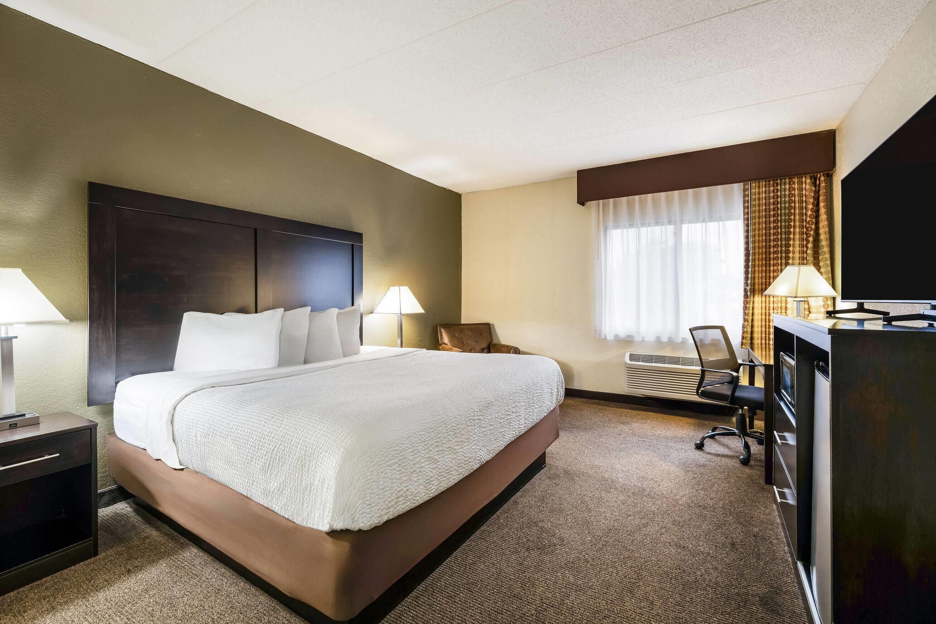 Et hotelværelse med en stor seng, sengeborde med lamper, et skrivebord med computer, et fjernsyn og et vindue med gardiner.