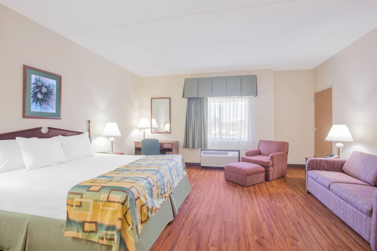 A hotel room with a bed, two armchairs, a sofa, a desk with a chair, a mirror, and a window with curtains.