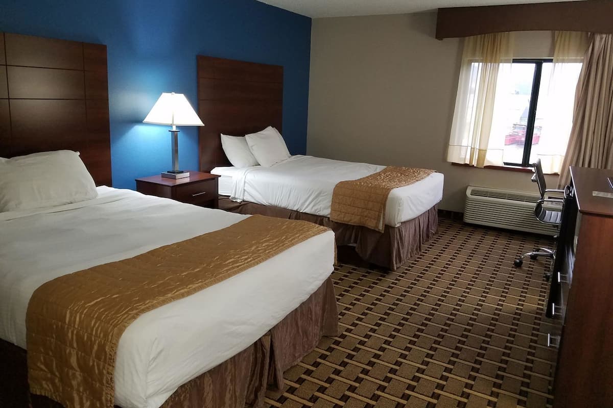 A hotel room with two beds, a nightstand with a lamp, an air conditioning unit, and a window with curtains.