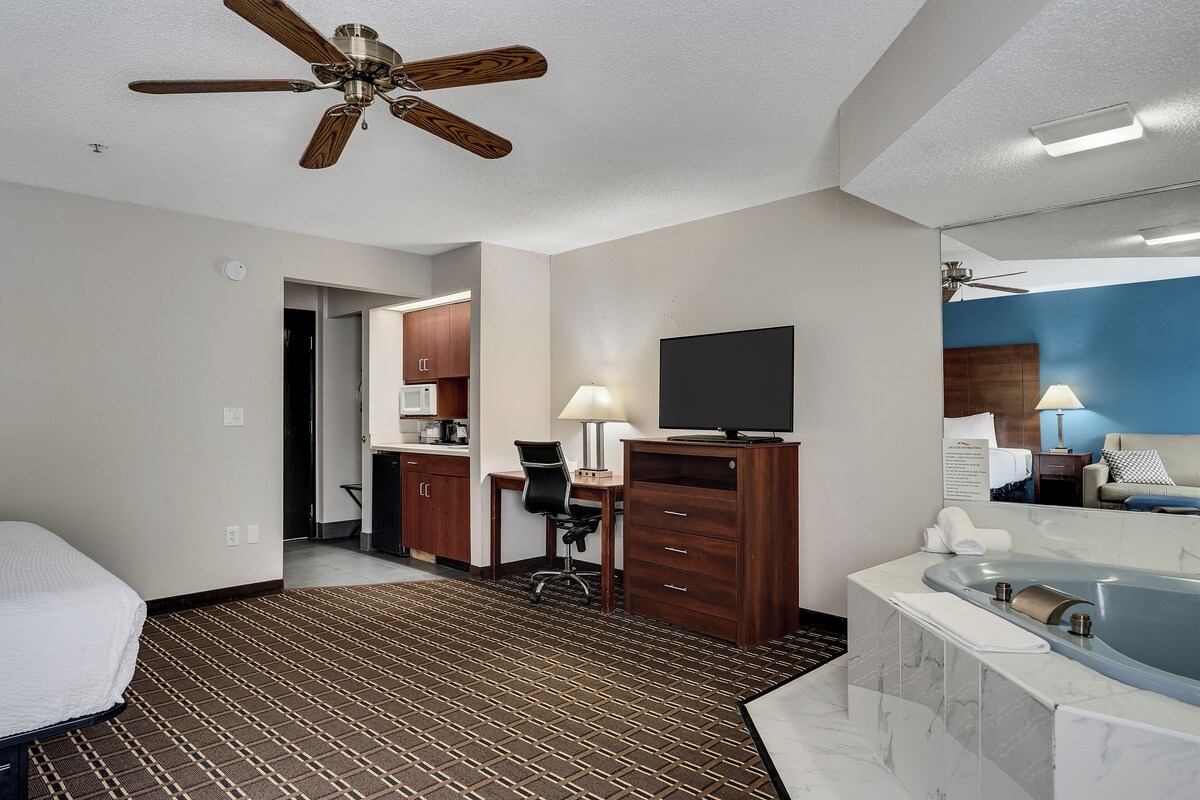 A hotel room with a bed, a desk with a chair, a desk lamp, a television, a bathroom with a bathtub, and a ceiling fan.