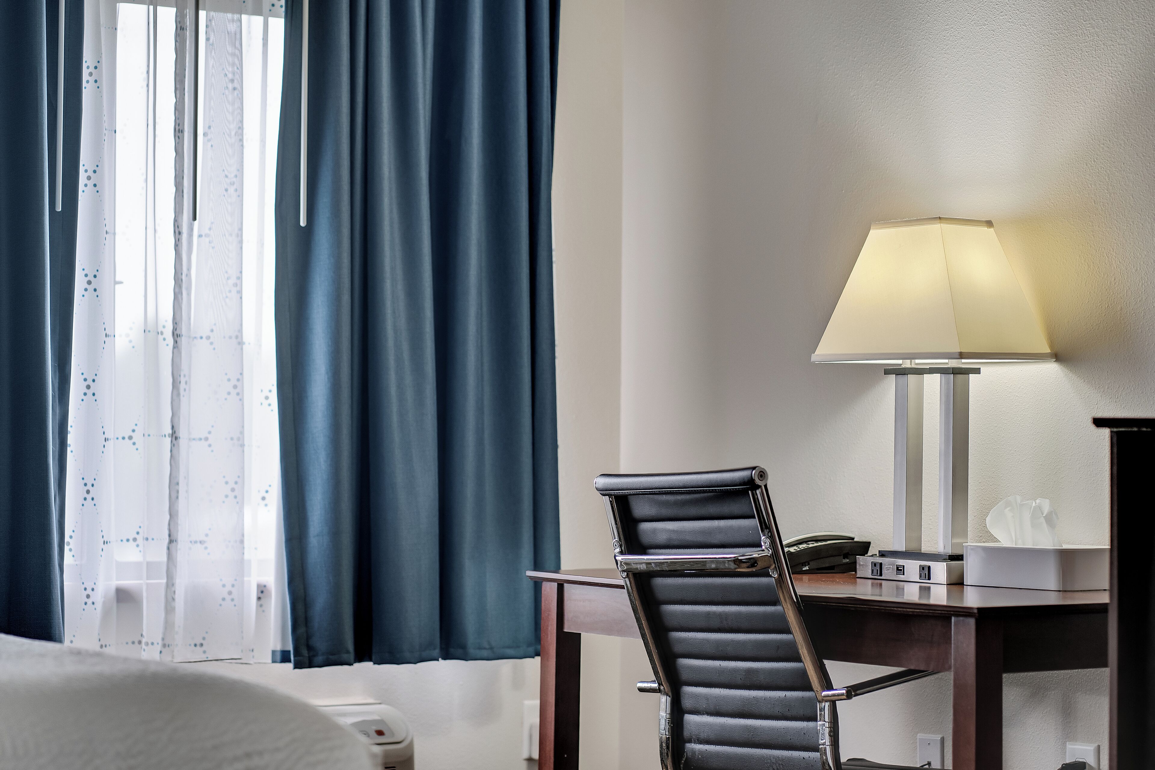 A hotel room with a bed, a desk with a lamp, a chair, and a window with curtains.