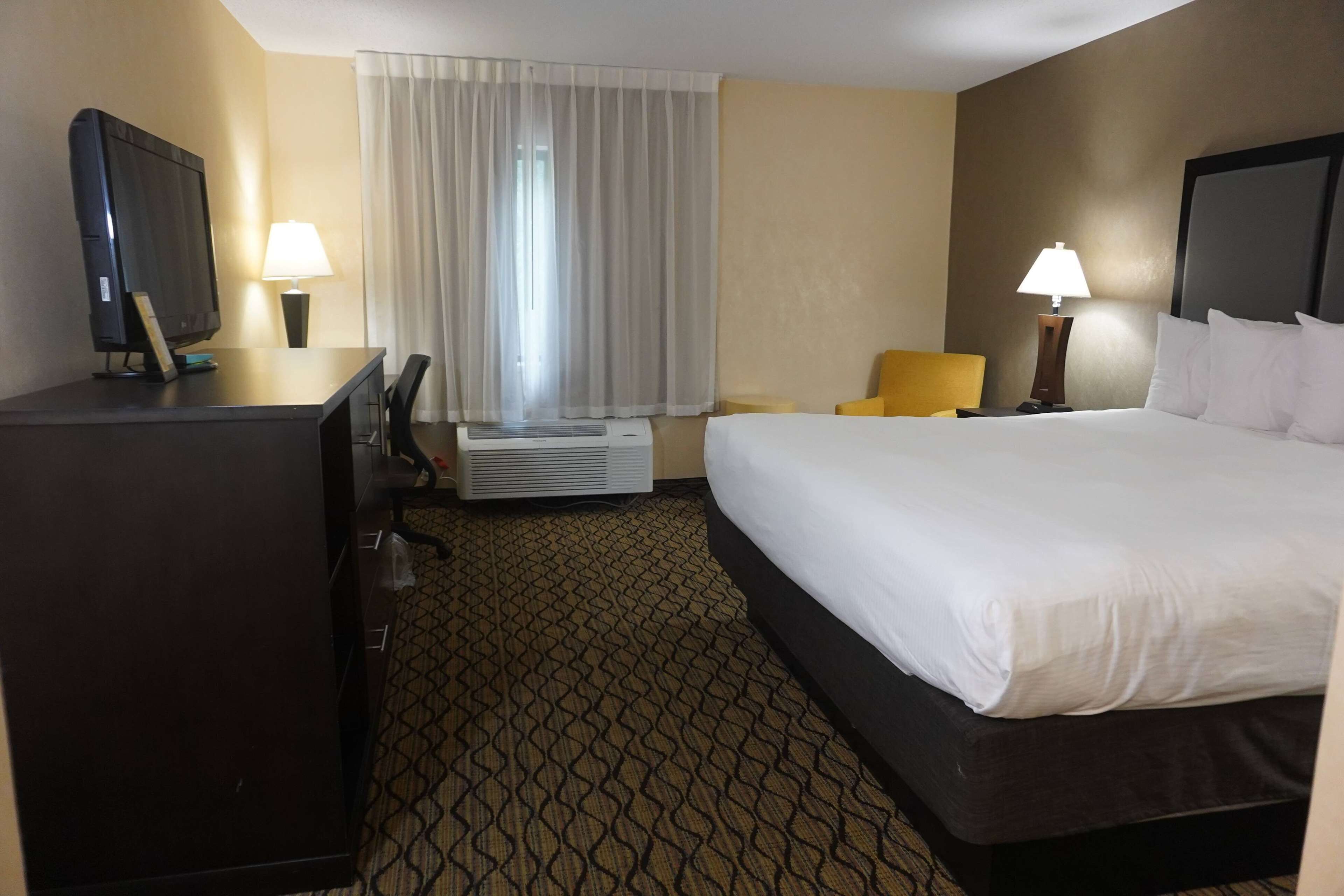Ein Hotelzimmer mit einem Bett, einem Schreibtisch mit Fernseher, einem Stuhl, einer Lampe und einem Fenster mit Vorhängen.
