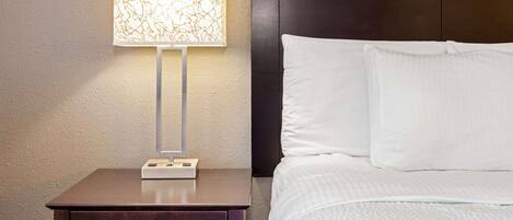 A neatly made bed with white linens, a bedside table with a lamp, and a dark headboard.