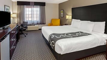 Quarto de hotel com uma cama grande, uma mesa de trabalho com uma TV, uma cadeira e uma janela com cortinas.