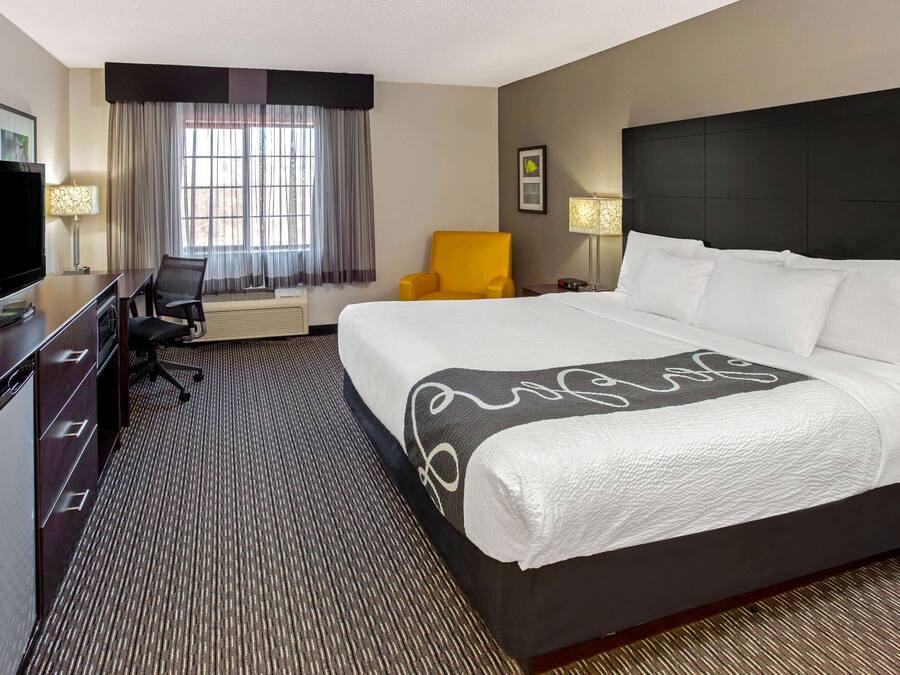 Quarto de hotel com uma cama grande, uma mesa de trabalho com uma TV, uma cadeira e uma janela com cortinas.