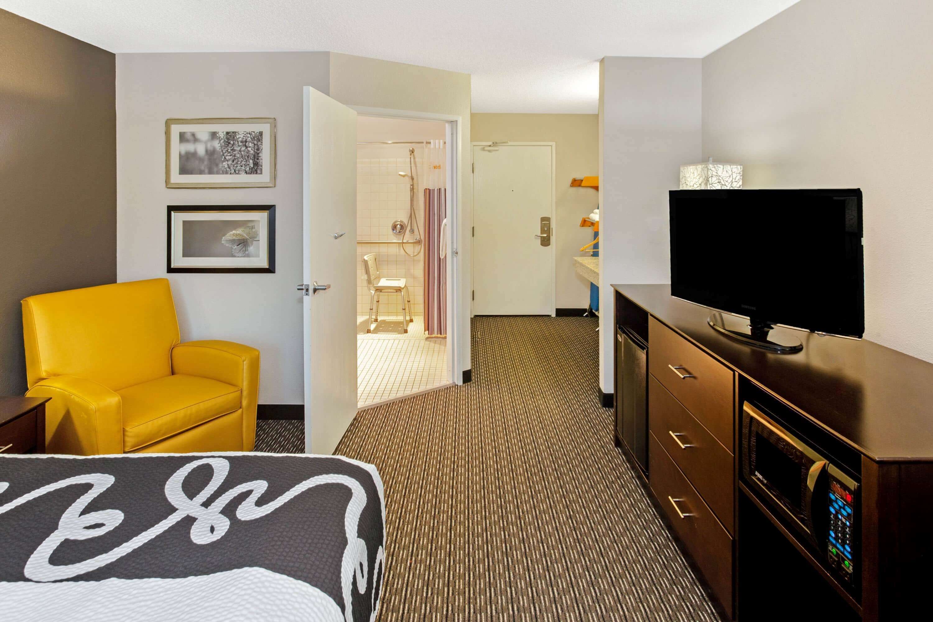 A hotel room with a bed, a television, a yellow armchair, a dresser, and a bathroom visible through an open door.