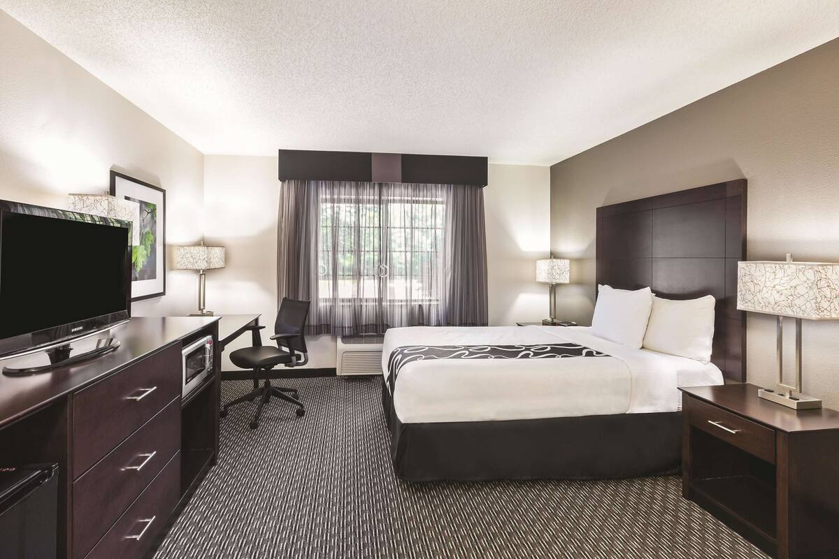 A hotel room with a bed, a TV, a desk, a chair, and a window with curtains.