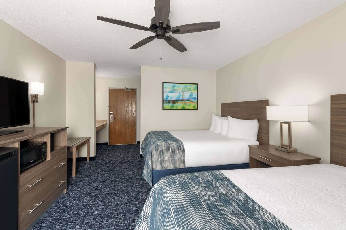Ein Hotelzimmer mit Bett, Schreibtisch mit Computer, Fernseher, Deckenventilator und einem Bild an der Wand.