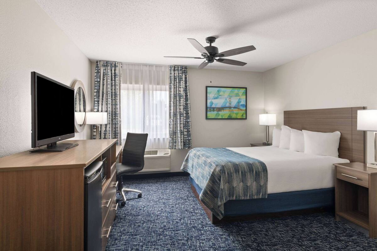 Ein Hotelzimmer mit einem Bett, einem Schreibtisch mit Fernseher, einem Sessel, einem Deckenventilator und einem Bild an der Wand.