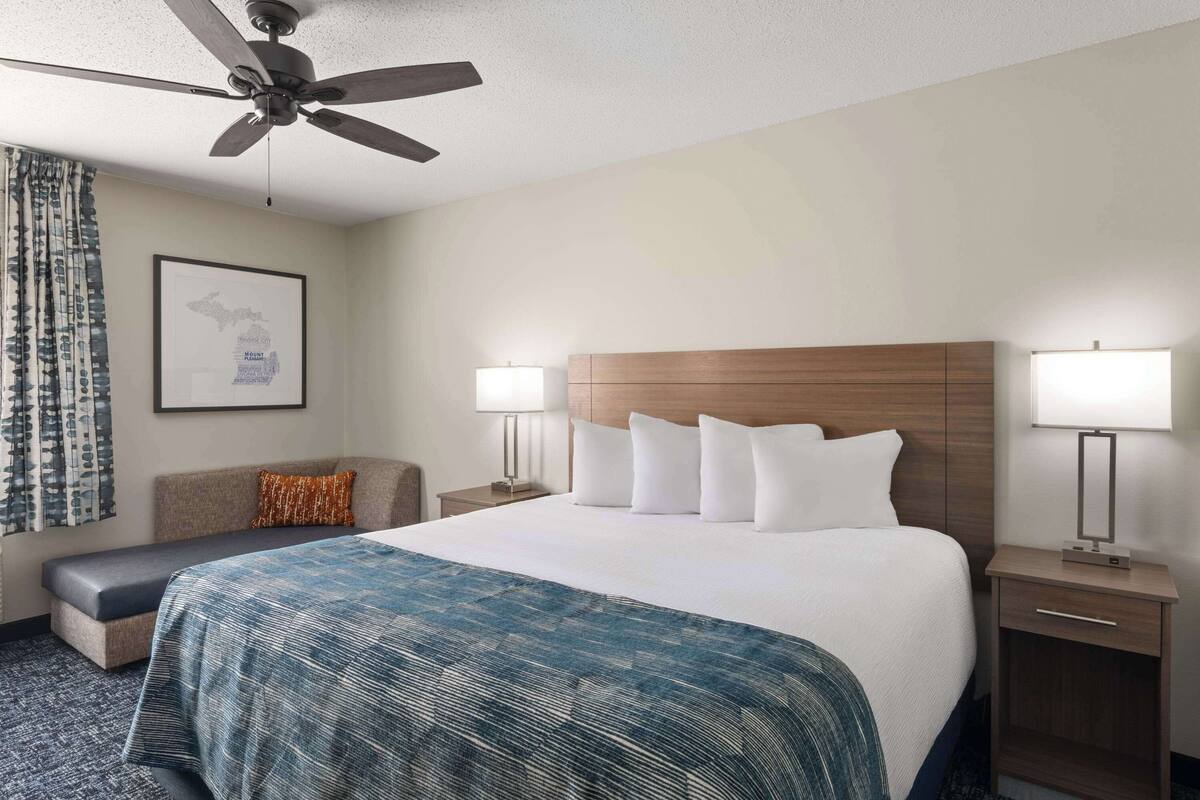Ein Hotelzimmer mit einem großen Bett, zwei Nachttischen mit Lampen, einem Deckenventilator, einer gerahmten Karte an der Wand und einem gemusterten Teppich.