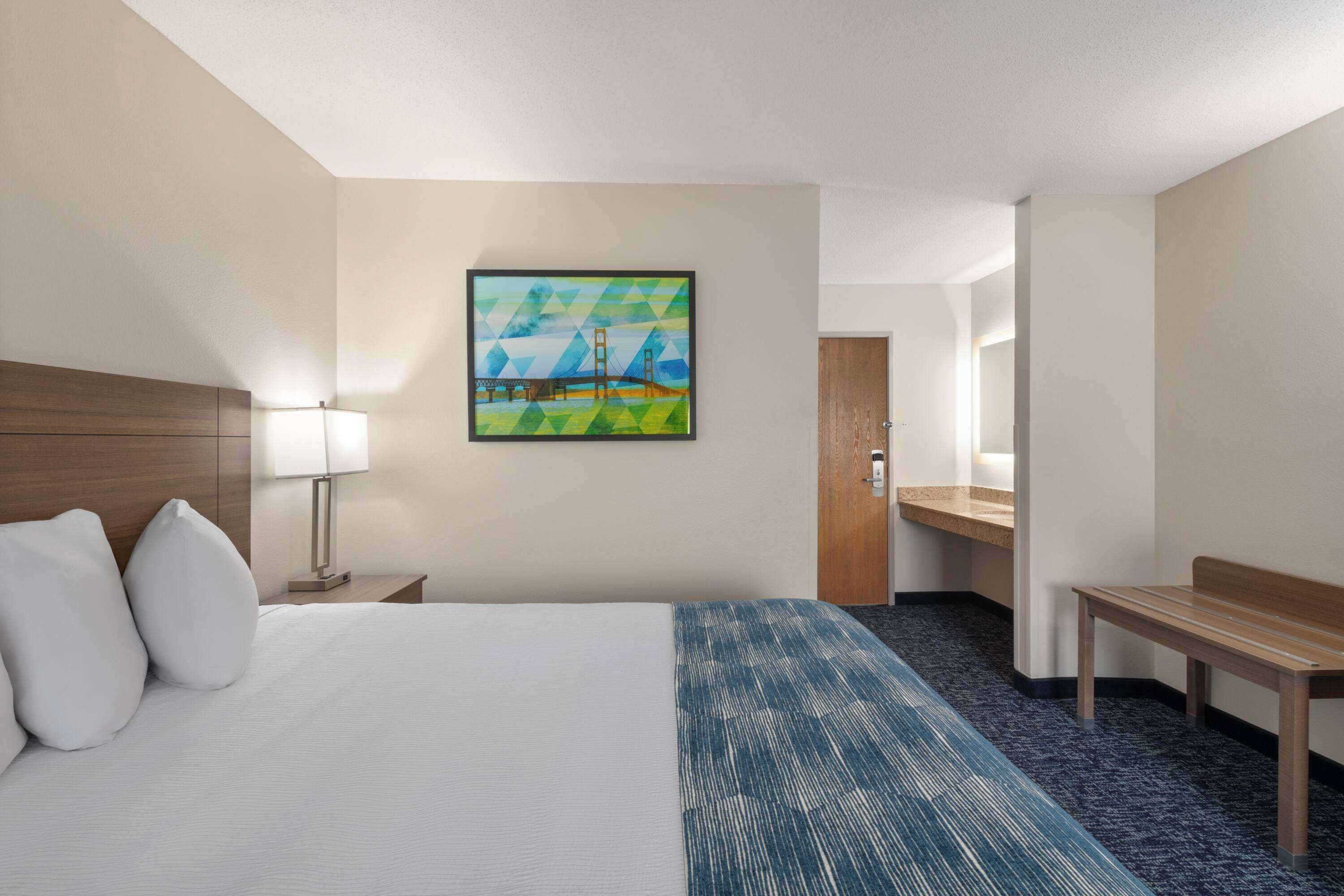 Una habitación de hotel con cama, escritorio, lámpara, un cuadro y un banco.
