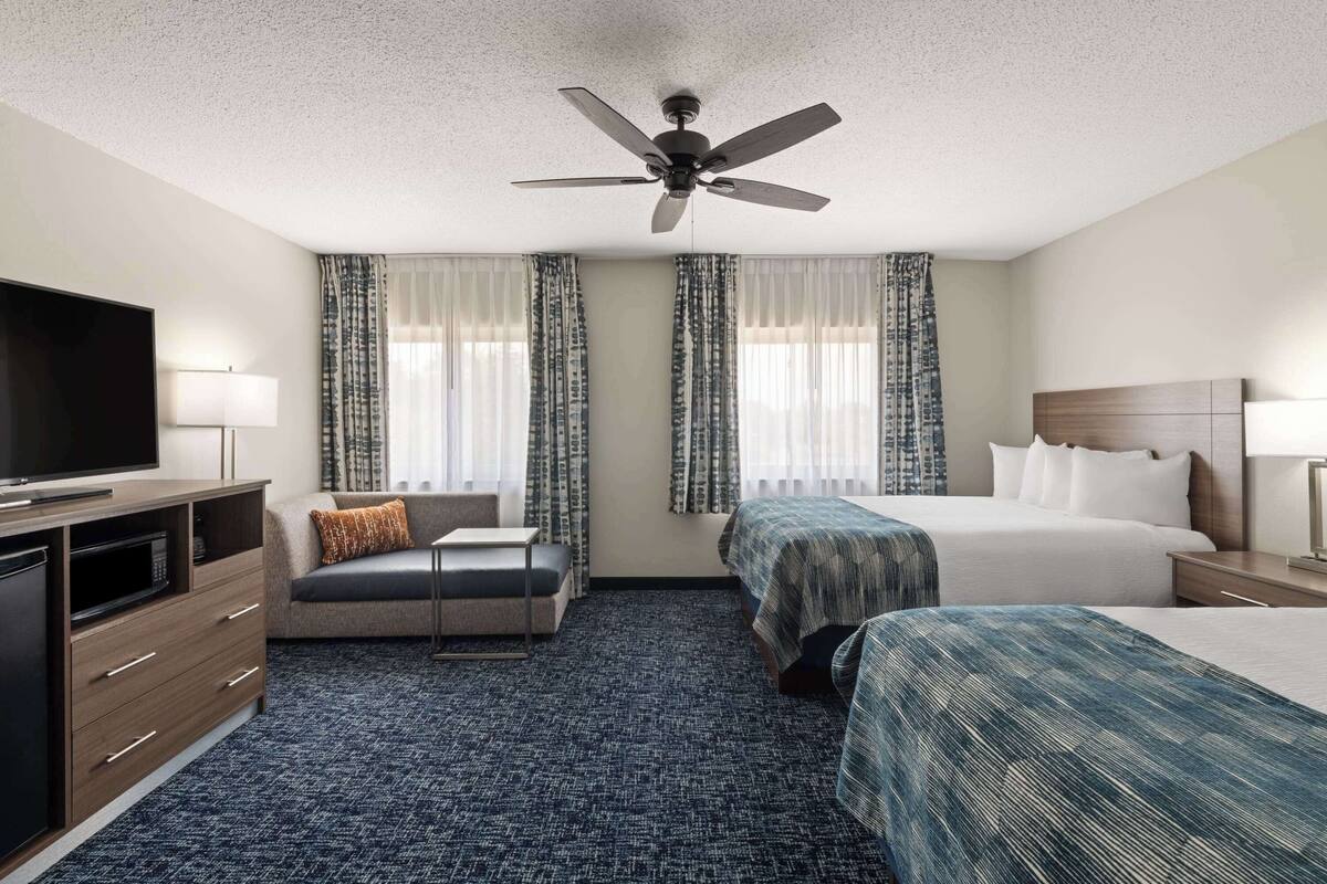 Ein Hotelzimmer mit Bett, Sofa, Fernseher, Schreibtisch und Deckenventilator.