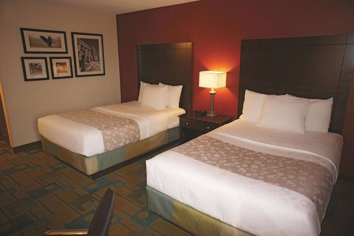 A hotel room with two beds, a nightstand with a lamp, a chair, and framed pictures on the wall.