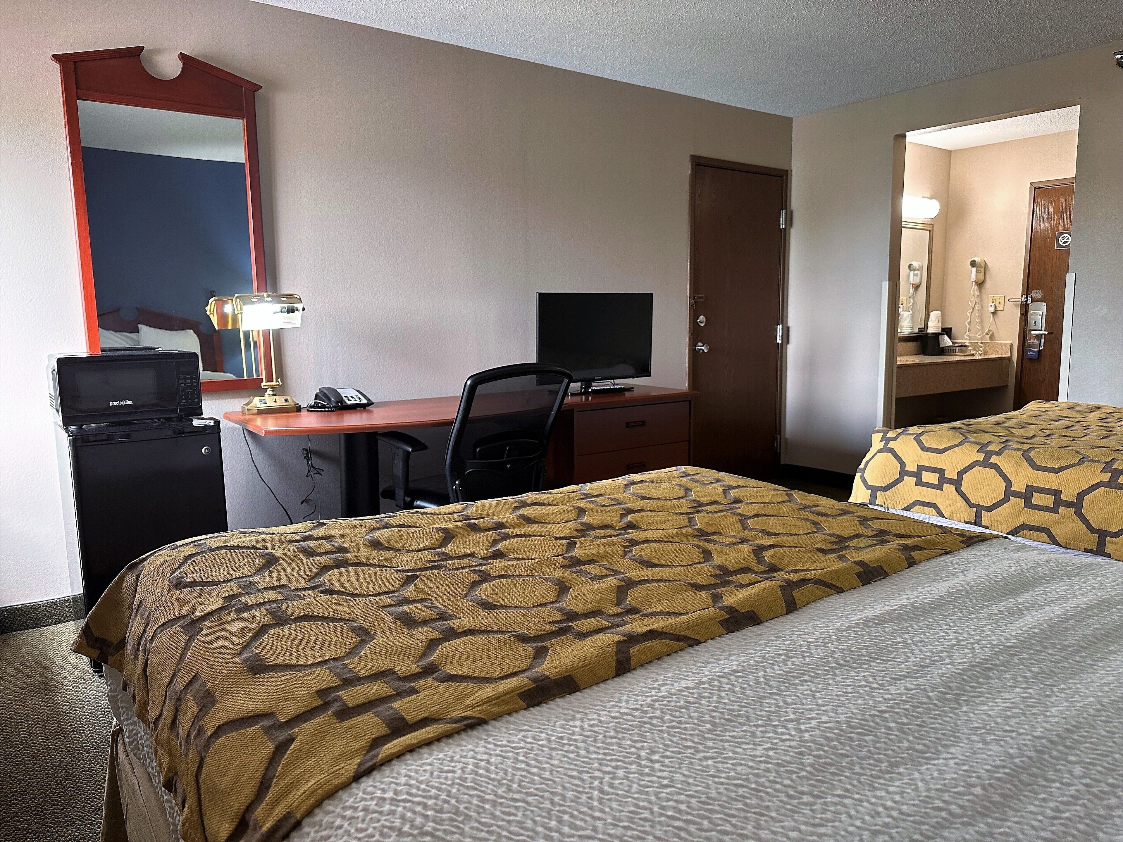 Hotelkamer met een bureau, een computer, een magnetron, twee bedden met behang en een spiegel, en een deur naar een badkamer.