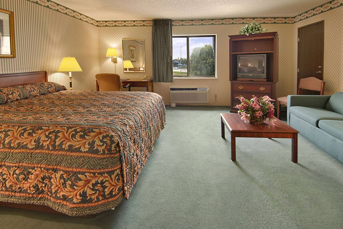 A hotel room with a large bed, a desk with a lamp, a television, a sofa, and a window with a view of trees.