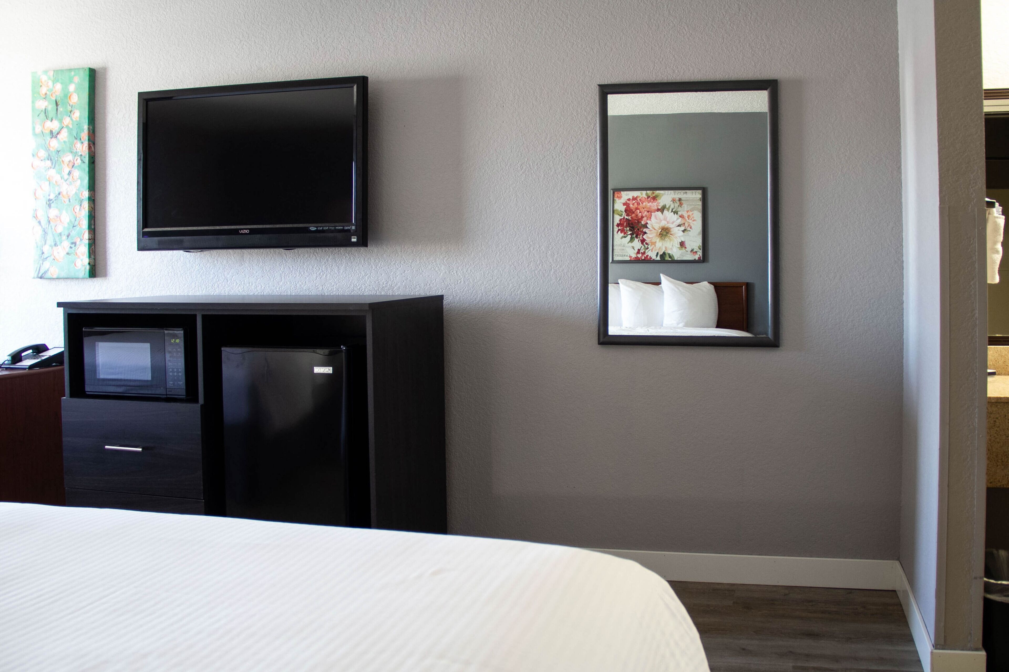 Habitación de hotel con cama, televisor montado en la pared, microondas, refrigerador y espejo que refleja parte del cuarto.