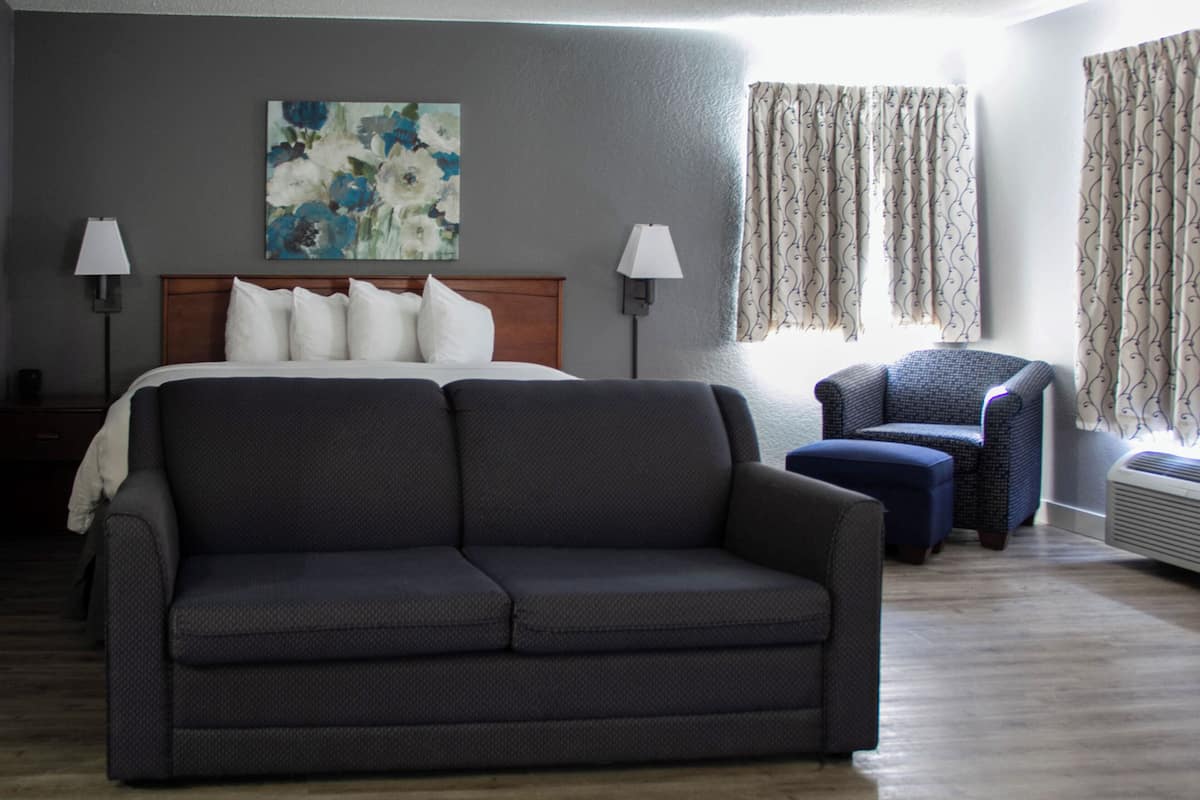 A hotel room with a large bed, a dark sofa, a chair, and a painting on the wall.