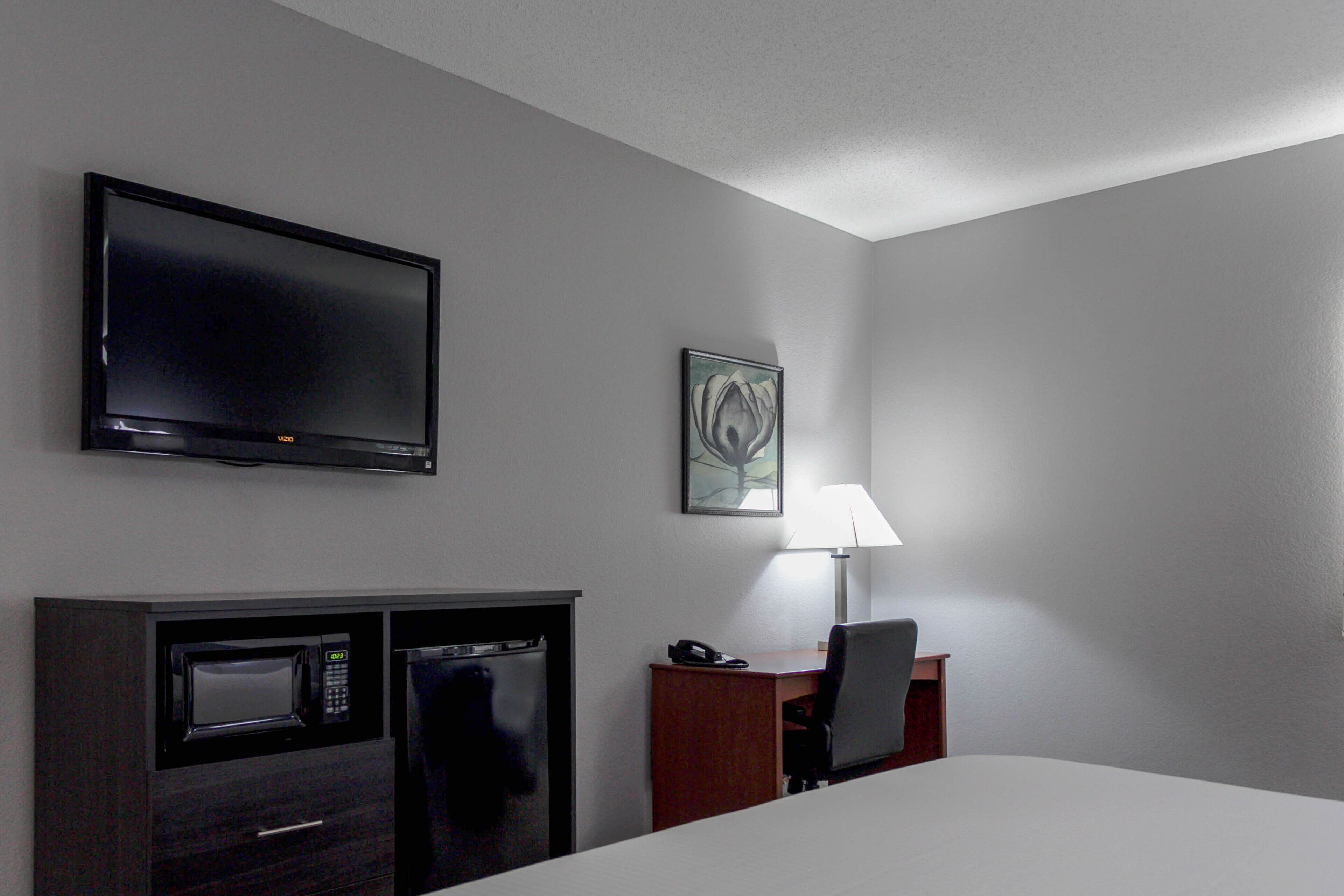 A hotel room with a flat-screen TV, a microwave, a refrigerator, a desk with a chair, a lamp, and a bed.
