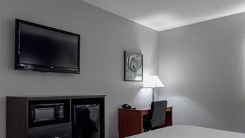 Ett hotellrum med en platt-TV, en mikrovågsugn, ett kylskåp, ett skrivbord med en stol, en lampa och en säng.