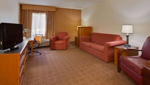 A hotel room with a television on a wooden desk, a red sofa, a matching armchair, a small table with a lamp, and a window with curtains. - La Quinta Inn & Suites by Wyndham Lakeland East (Lakeland)